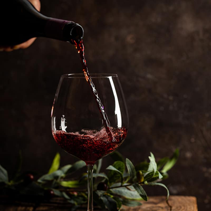 Vin rouge versé dans un verre élégant : dégustation chic Vin rouge versé dans un verre à pied sur fond sombre avec branche d'olivier et éclairage tamisé.